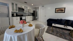 A modern kitchen with white cabinets, a microwave, and a refrigerator. There is a dining area with a table set for two, a sofa, and a small table with a remote control. - Bed & Breakfast Coconut Grove Miami (Miami)