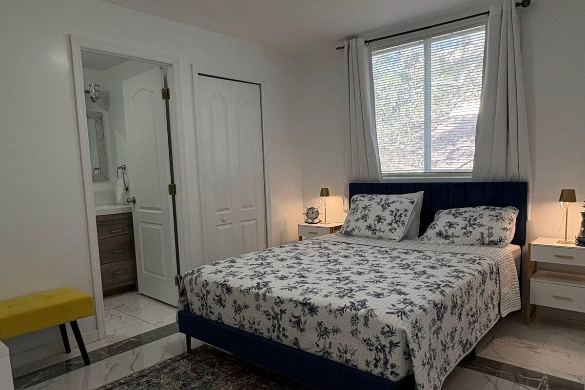 A bedroom with a bed, bedside tables, a nightstand, a window with curtains, a door, and a bathroom visible through an open door.