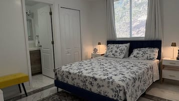 Un dormitorio con cama, mesitas de noche, una ventana con cortinas, una puerta y un baño visible a través de una puerta abierta.