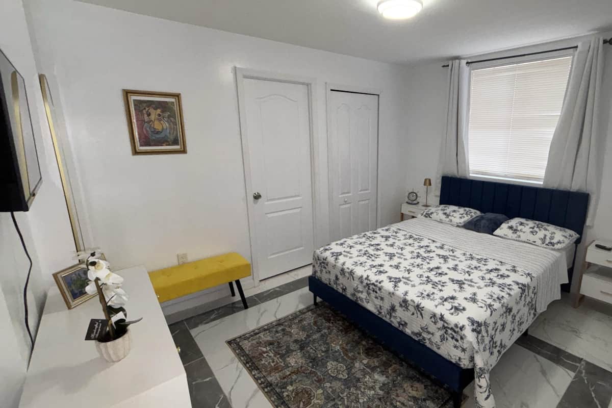 A bedroom with a bed, bedside tables, a bench, a rug, a window with curtains, and a door.