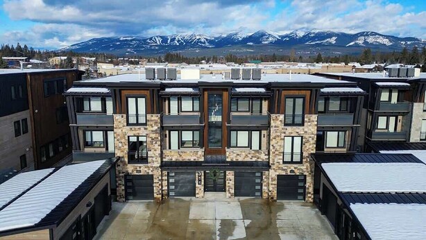 A multi-story residential building with a central entrance, garage doors, and a view of snow-capped mountains in the background. - Montana Magnificence Studio (Whitefish)