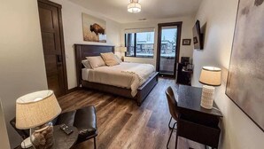 A compact hotel room with a bed, a desk, a chair, a lamp, and a large window offering a view. - Montana Magnificence Studio (Whitefish)