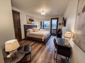 A compact hotel room with a bed, a desk, a chair, a lamp, and a large window offering a view. - Montana Magnificence Studio (Whitefish)