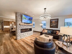 A modern living room with a stone fireplace, a flat-screen TV mounted on a stone wall, a black leather sofa, and a chandelier. - Montana Magnificence Chalet (Whitefish)