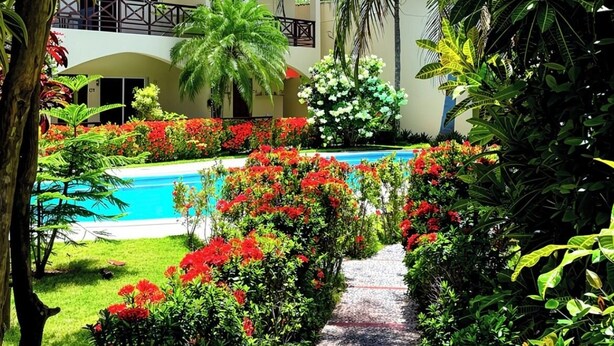 Un sentier mène à une piscine entourée d’une végétation luxuriante et à un bâtiment doté de balcons.