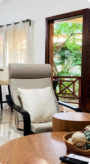 A room with a beige armchair, a small wooden table, a remote control, and a wicker basket. - Relax & Explore: Cozy 2BR with Pool Parking & WiFi (Las Terrenas)