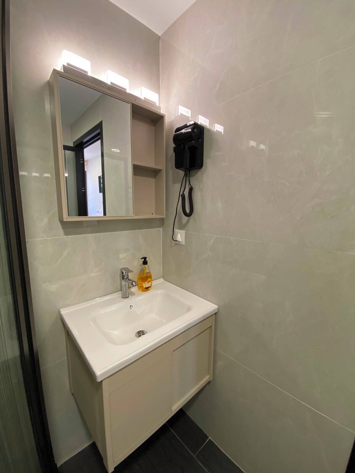 Un baño con lavamanos blanco, un espejo, un secador de pelo montado en la pared y una botella de jabón.