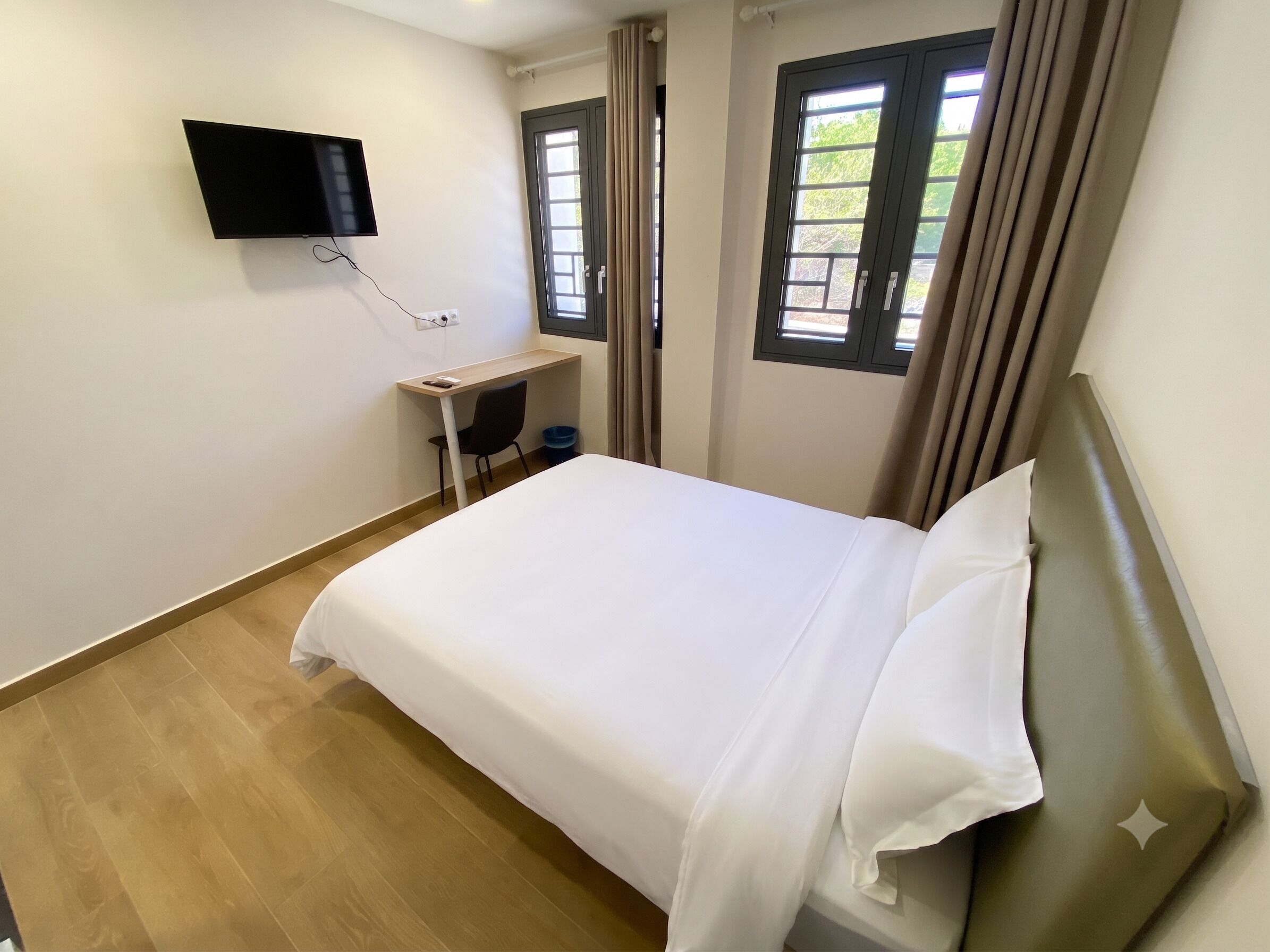 Un dormitorio con cama, escritorio, silla, televisor montado en la pared y dos ventanas con cortinas.