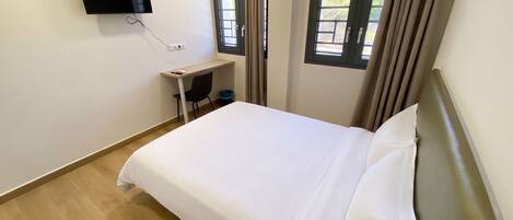 A bedroom with a bed, a desk, a chair, a television mounted on the wall, and two windows with curtains.