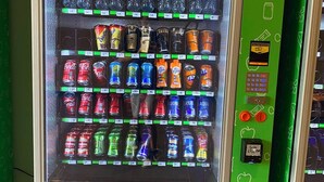 A vending machine with a green exterior, a push button, and various beverage cans and bottles.