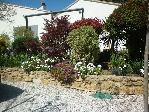 Ein Garten mit vielfältigen Pflanzen und Blumen, eine Steinmauer und ein Gebäude mit heller Fassade und Schornstein.