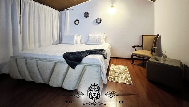 Quarto com teto de madeira, cama com edredom branco, uma cadeira, uma mesinha e luminária fixada na parede.