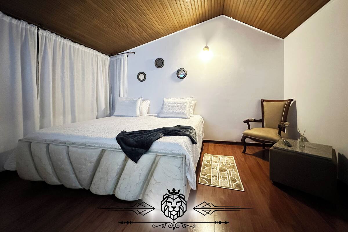 Ein Schlafzimmer mit Holzbalkendecke, einem Bett mit weißem Duvet, einem Sessel, einem kleinen Tisch und einer Wandleuchte.