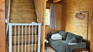 Interior de una cabaña de madera con escaleras, un sofá, una lámpara de pie y una ventana con cortinas.