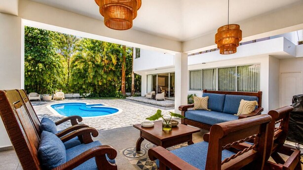 Un área de piscina con piscina azul, mobiliario de madera y vistas a un jardín exuberante.