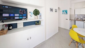 Una habitación moderna con televisor de pantalla plana, una zona de comedor con sillas amarillas y una cocina con armarios blancos.