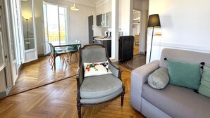 A spacious living room with a sofa, a dining table with chairs, a kitchen area, and a large window. - Balcon Palais Excelsior Grimaldi (Nice)