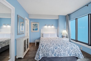 A bedroom with a bed, bedside tables, a dresser, a large window, and wall art. - A Coastal Paradise - Beachfront - Gym - Pool (Myrtle Beach)