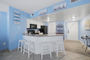 A kitchen with a central island, white bar stools, a microwave, a refrigerator, and a sink. - A Coastal Paradise - Beachfront - Gym - Pool (Myrtle Beach)