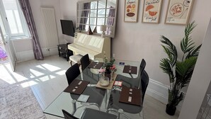 A dining area with a glass table, chairs, a piano, and framed artwork on the wall. - Red House  (Guildford)
