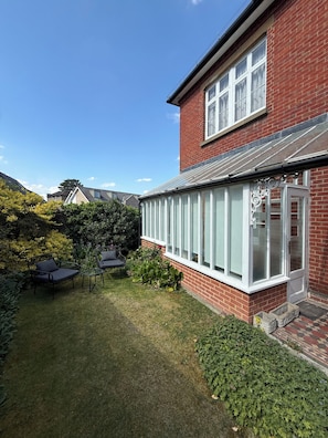 A residential property with a brick exterior, a conservatory with glass doors, a lawn, and outdoor furniture.