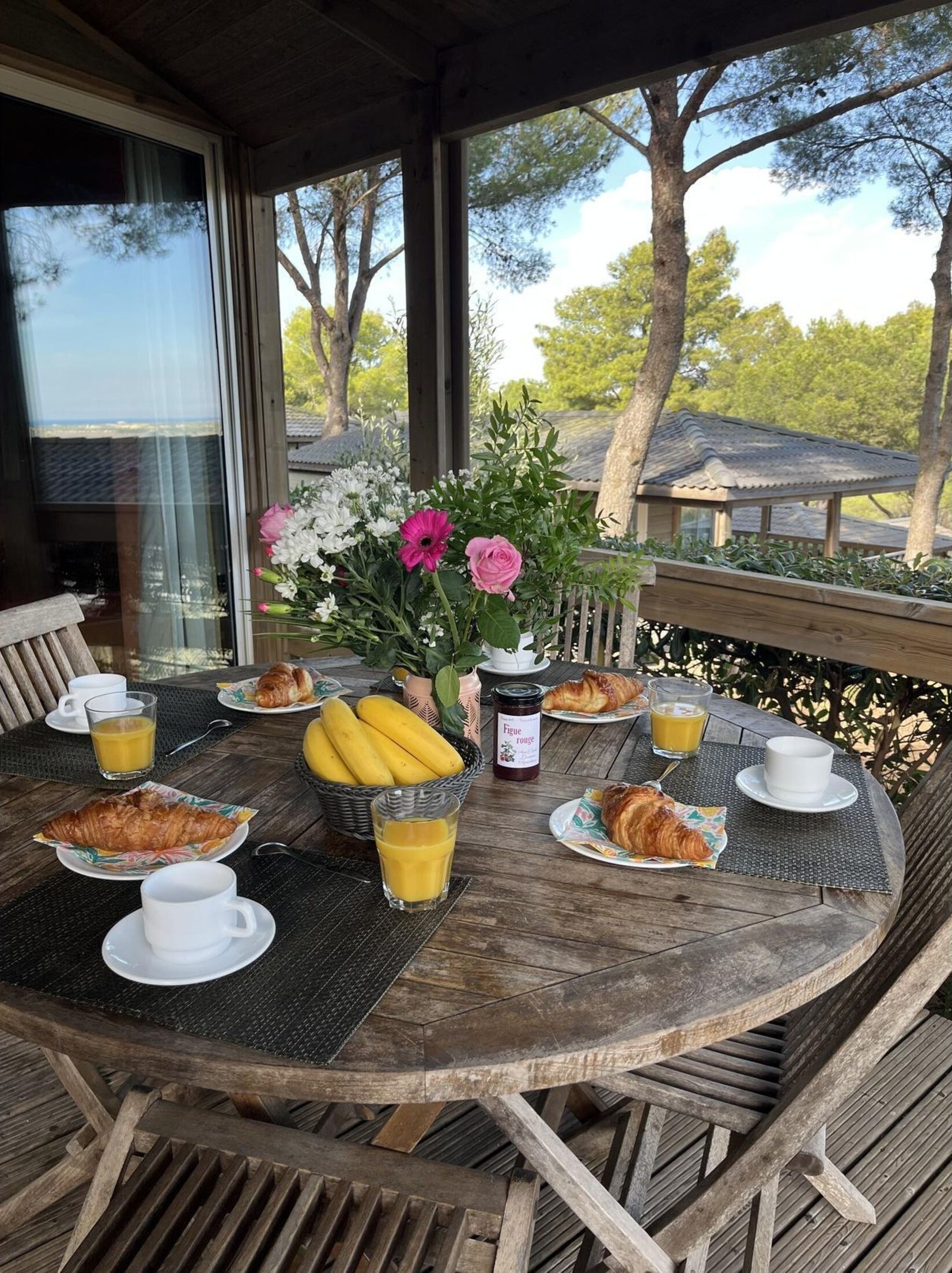 A round wooden table with breakfast items, including croissants, bananas, and juice, set outdoors with a view of trees and a building in the background.