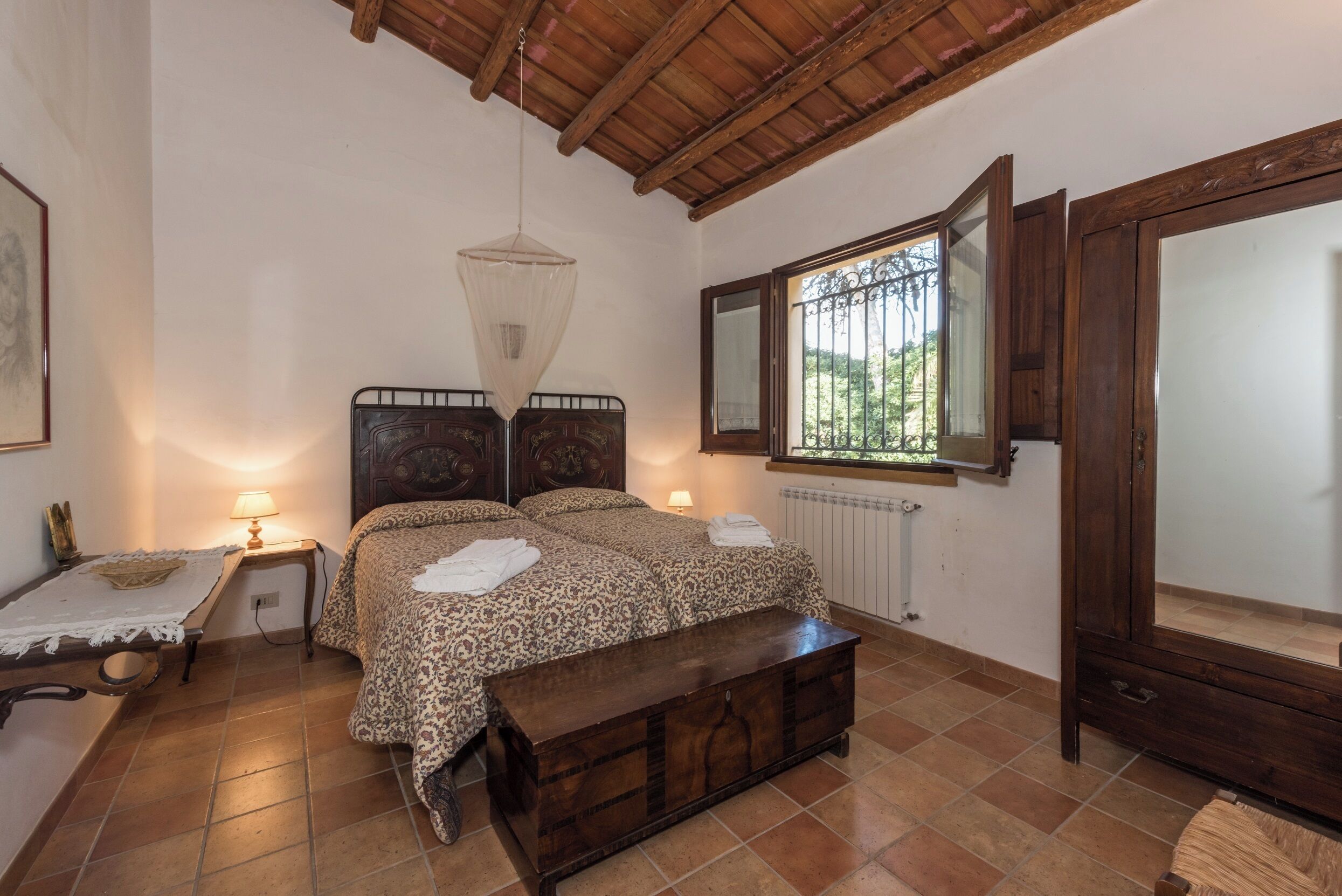 A bedroom with a bed, bedside lamps, a wooden bench, a window with a view, and a radiator.