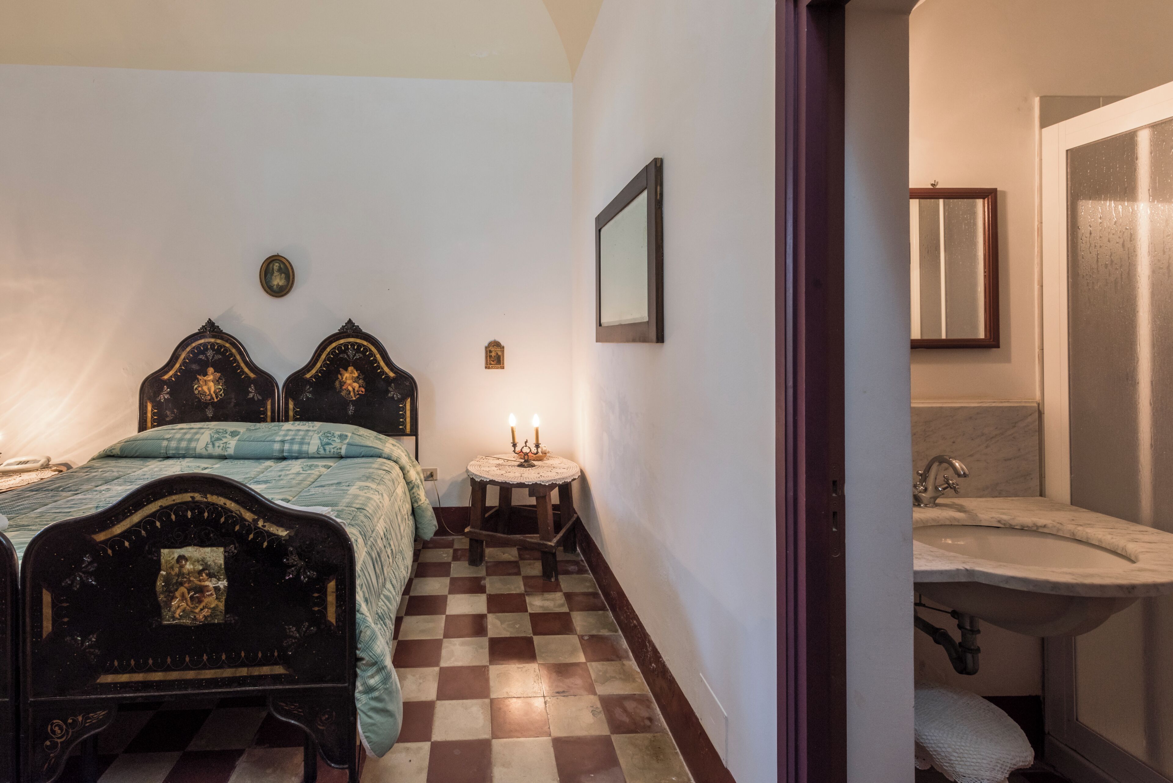 A bedroom with a checkered floor, a bed with ornate headboard, a bedside table with a lamp, and a small round table with candles.