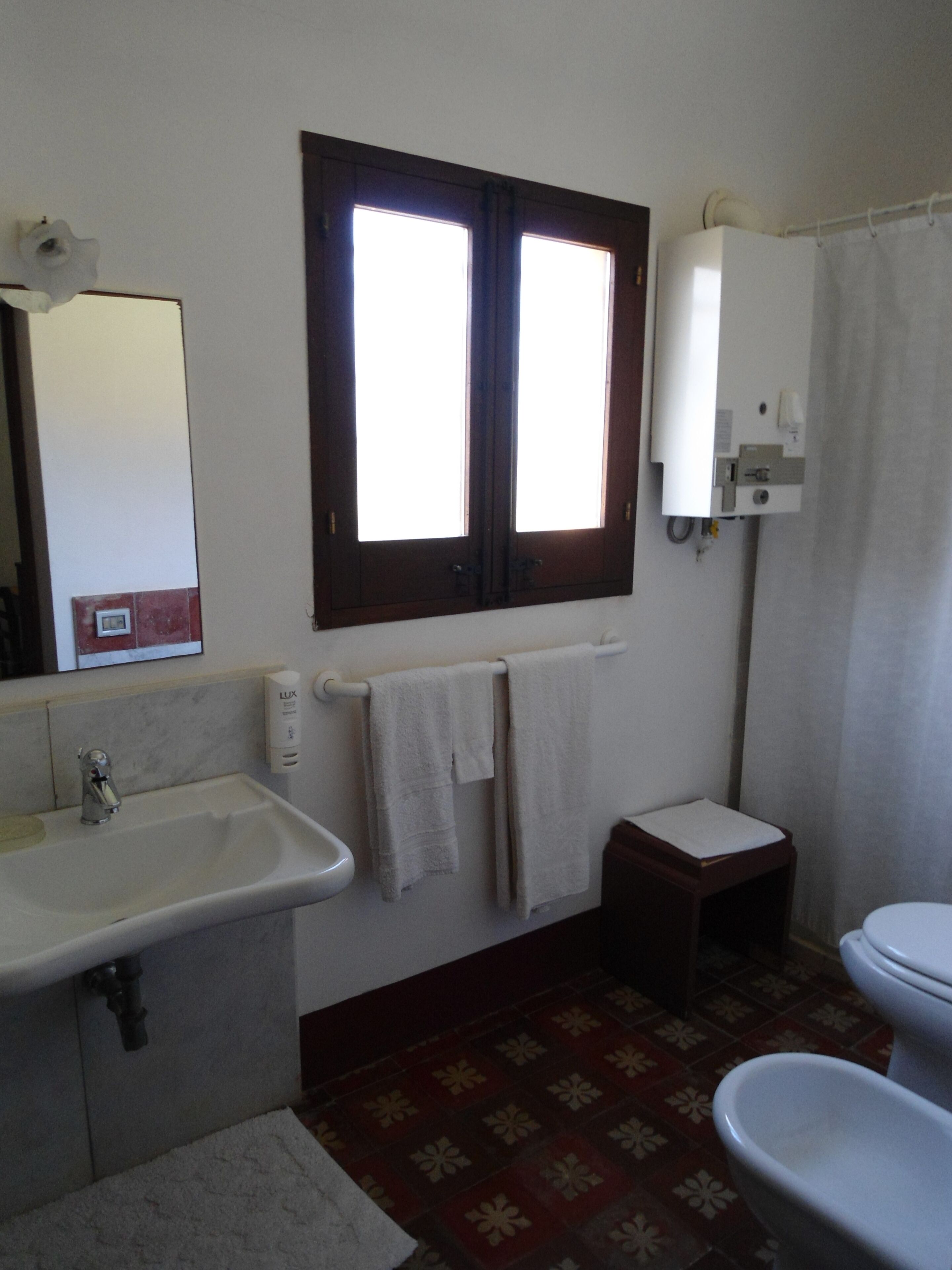 A bathroom with a sink, mirror, window, towel rack, toilet, and patterned floor.