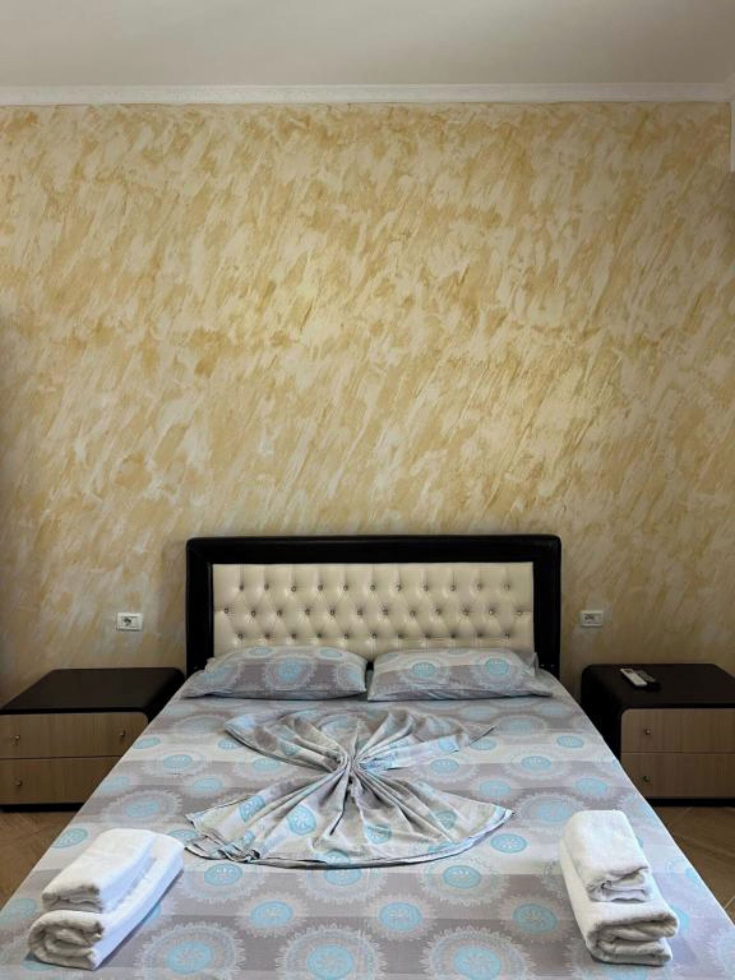 A bedroom with a tufted headboard, a patterned bedspread, two bedside tables, and folded towels.