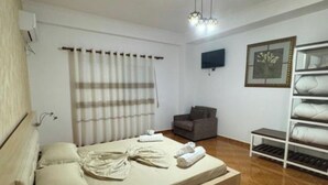 Ein Schlafzimmer mit Bett, Sessel, Fernseher, Ablage mit Handtüchern und Fenster mit Vorhängen.