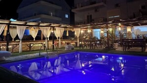 Ein Poolbereich bei Nacht mit einem blau beleuchteten Pool, Sitzgelegenheiten im Freien und einem Gebäude im Hintergrund.