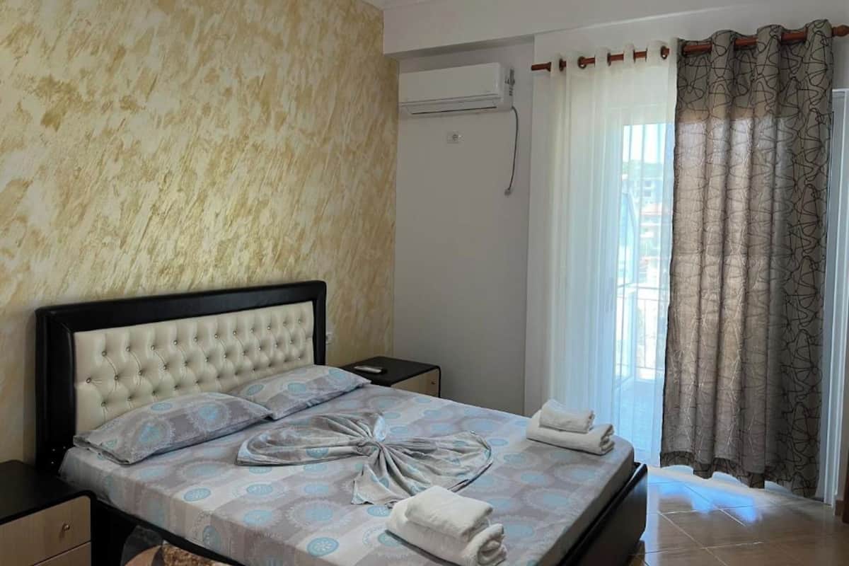 A hotel room with a bed, a nightstand, an air conditioning unit, a window with curtains, and a view of the outside.