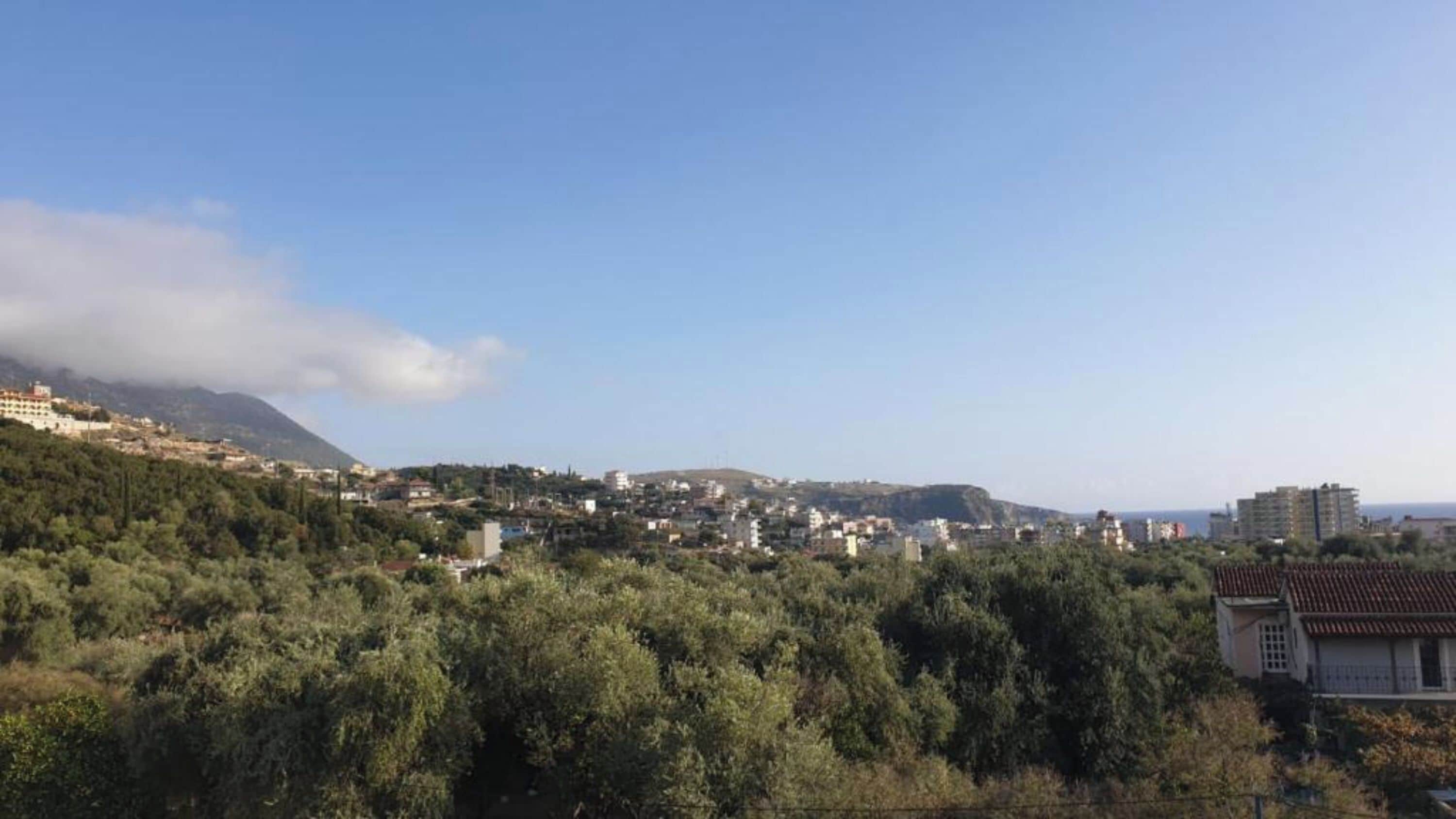 A town with buildings on a hillside, a mountain in the background, and dense greenery in the foreground.