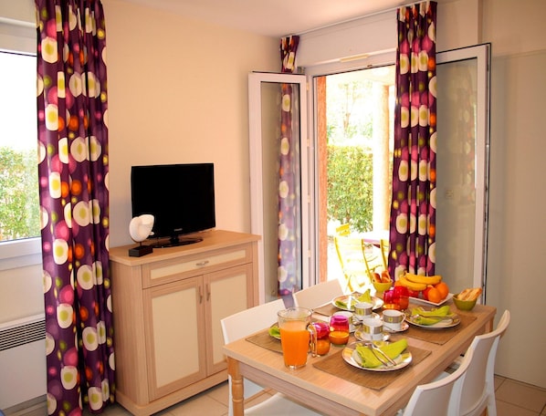 A room with a TV on a wooden cabinet, a dining table with breakfast items, and curtains with a circular pattern. - Spacious Coastal Retreat for 6 (Saint-Cyprien)