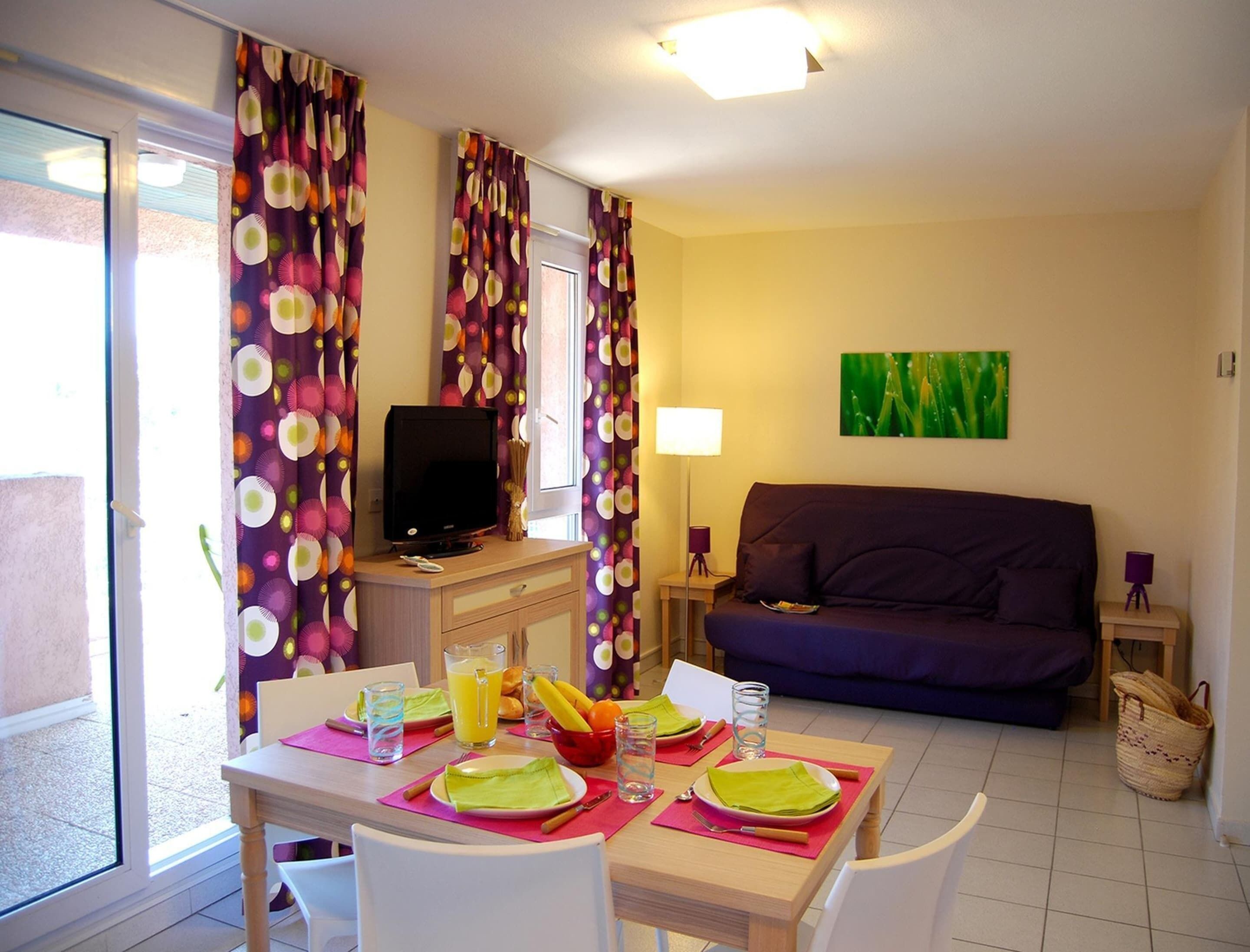 Un salon avec un canapé, un téléviseur posé sur un meuble, une table à manger dressée avec des assiettes, des verres et une carafe de jus.