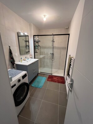 Un bany compacte amb rentadora, dutxa amb envàs de vidre, un lavabo amb mirall i un suport per a tovalloles.