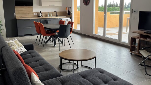 Ein modernes Wohnzimmer mit grauem Sofa, einem Esstisch mit Stühlen, einer Küchenzeile und einem Balkon mit gelber Wand.