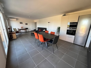 Une cuisine moderne avec des carreaux gris, une table à manger avec des chaises orange, un canapé noir et un réfrigérateur en acier inoxydable.
