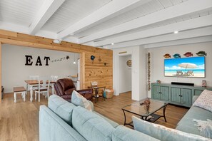 Ein modernes Wohnzimmer mit Holzbalkendecke, einem Fernseher, der eine Strandansicht zeigt, einem Essbereich mit einer Wandtafel, auf der 'EAT' steht, und einer gemütlichen Sitzecke mit einem blauen Sofa.