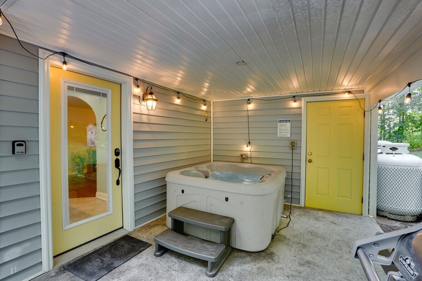 A covered outdoor area with a hot tub, a bench, and a door leading to a house.