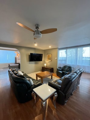 A spacious living room with a ceiling fan, a flat-screen TV, leather sofas, a coffee table, and large windows.