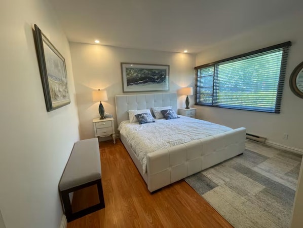 A bedroom with a bed, bedside tables, a bench, a window with blinds, and a framed picture on the wall.