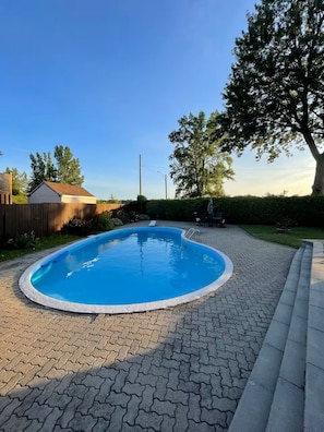 A rectangular swimming pool with a tiled deck, surrounded by a wooden fence and a tree.