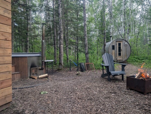 A rustic cabin with a wood-burning stove, a wooden chair, and a small shed in a forested area.