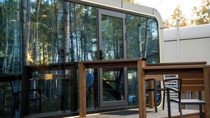 A modern building with large glass windows reflecting trees, a wooden bench, and a staircase leading to an upper level. - The Spruce Bliss Pod (Lac du Bonnet)