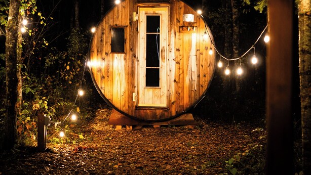 A wooden cabin with a door, windows, and a small porch area, surrounded by trees and string lights. - The Spruce Bliss Pod (Lac du Bonnet)