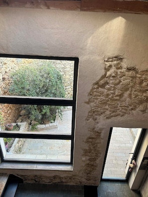 A room with a large window, a stone wall, and a view of an outdoor garden area. - L'Amandine - Renovated barn with Jacuzzi (Puisserguier)