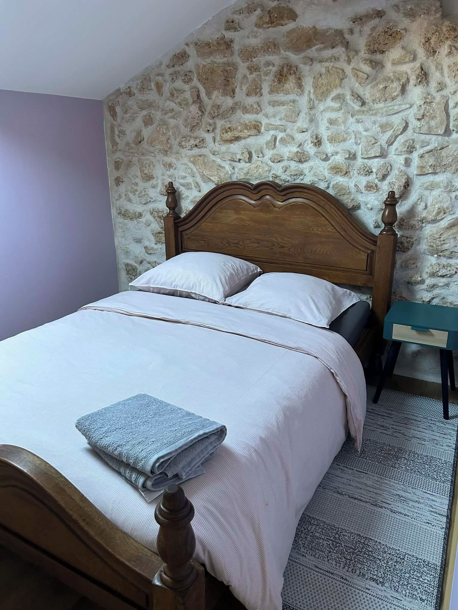 A bedroom with a stone wall, a wooden bed with a headboard, two pillows, a folded towel, and a small bedside table.