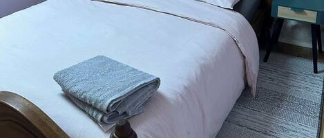 A bedroom with a stone wall, a wooden bed with a headboard, two pillows, a folded towel, and a small bedside table.